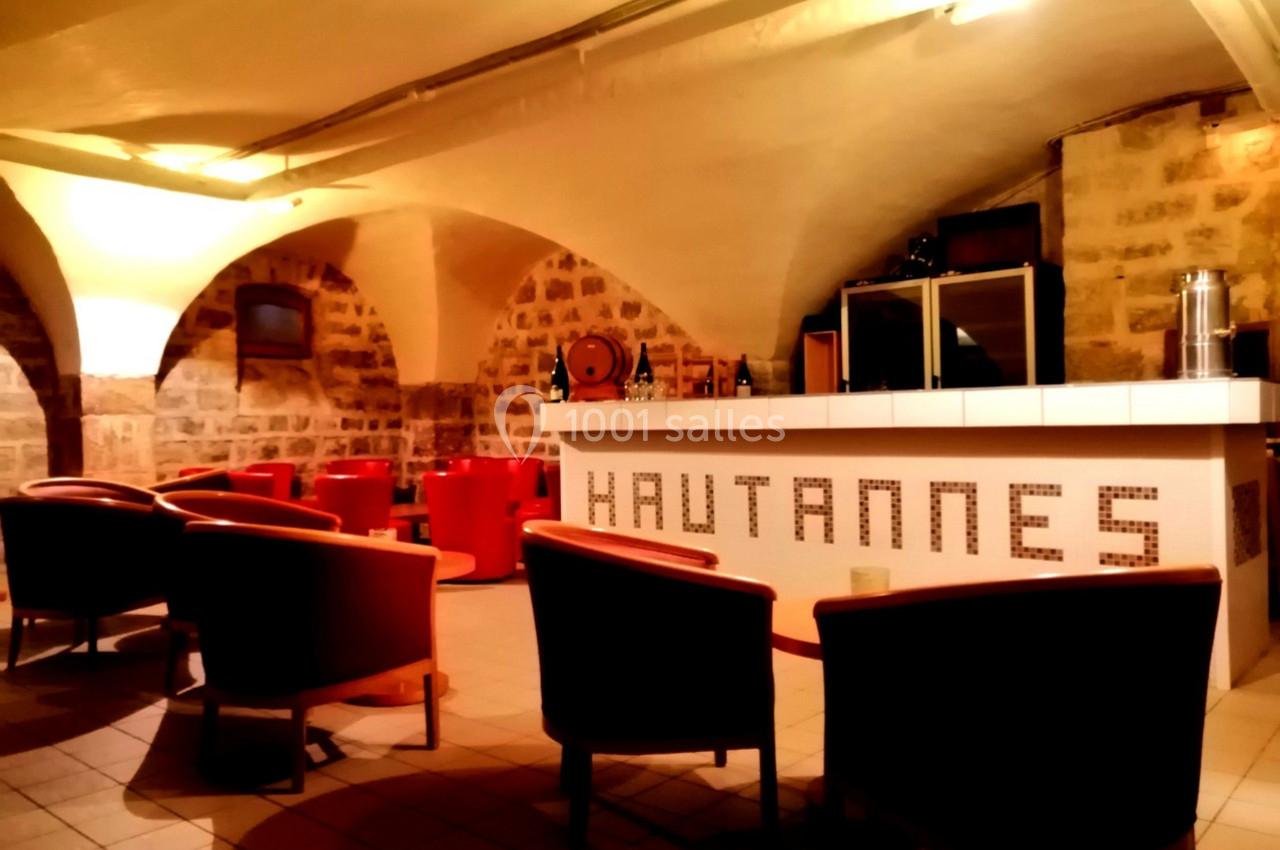 Salle voûtée avec des murs en pierre, un bar blanc marqué ’Hautannes’ et des fauteuils rouges disposés autour de tables.