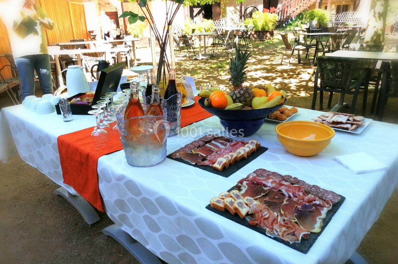 Table dressée en extérieur avec charcuterie, fruits, boissons et vaisselle, sous un espace ombragé.