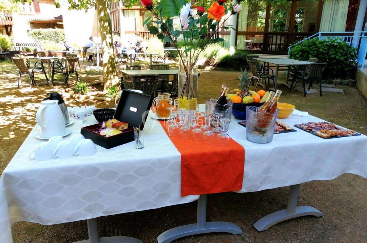 Table dressée en extérieur avec boissons, fruits, viennoiseries et verres, sous des arbres dans un cadre convivial.