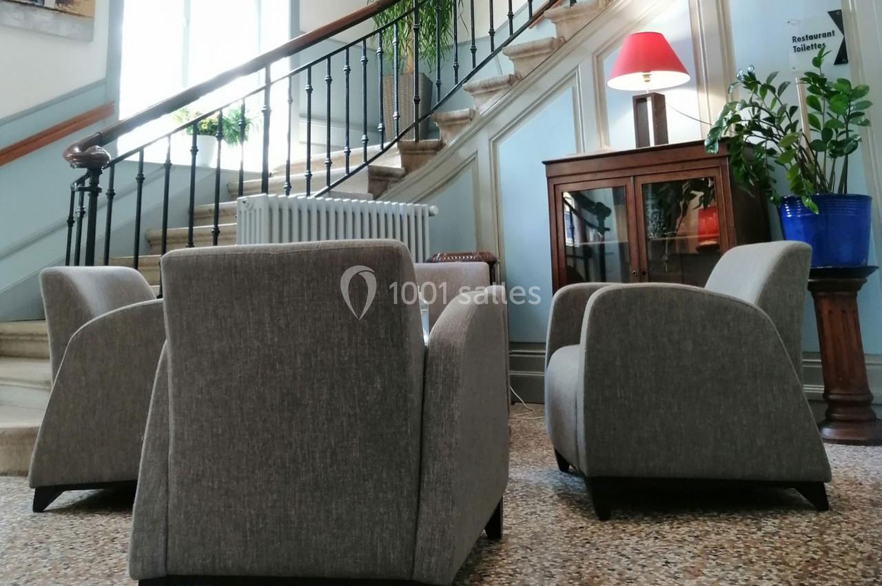 Trois fauteuils gris disposés autour d'une table basse dans un hall avec escalier en colimaçon et plantes décoratives.