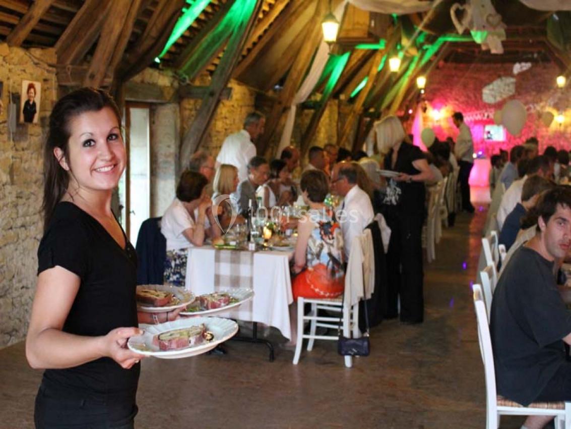 Une serveuse souriante apporte des plats dans une salle de réception rustique avec des convives attablés.