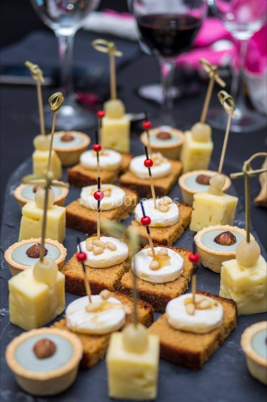 Assortiment de bouchées apéritives sur un plateau, comprenant fromage, tartelettes et gâteaux, avec des verres en arrière…