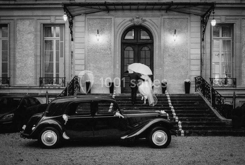 Un couple sous un parapluie monte les marches d'un bâtiment élégant, avec une voiture ancienne au premier plan.