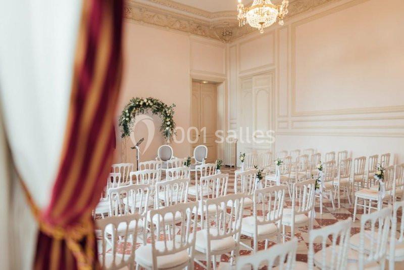 Salle de cérémonie lumineuse avec des chaises blanches alignées et une arche florale décorative au fond.