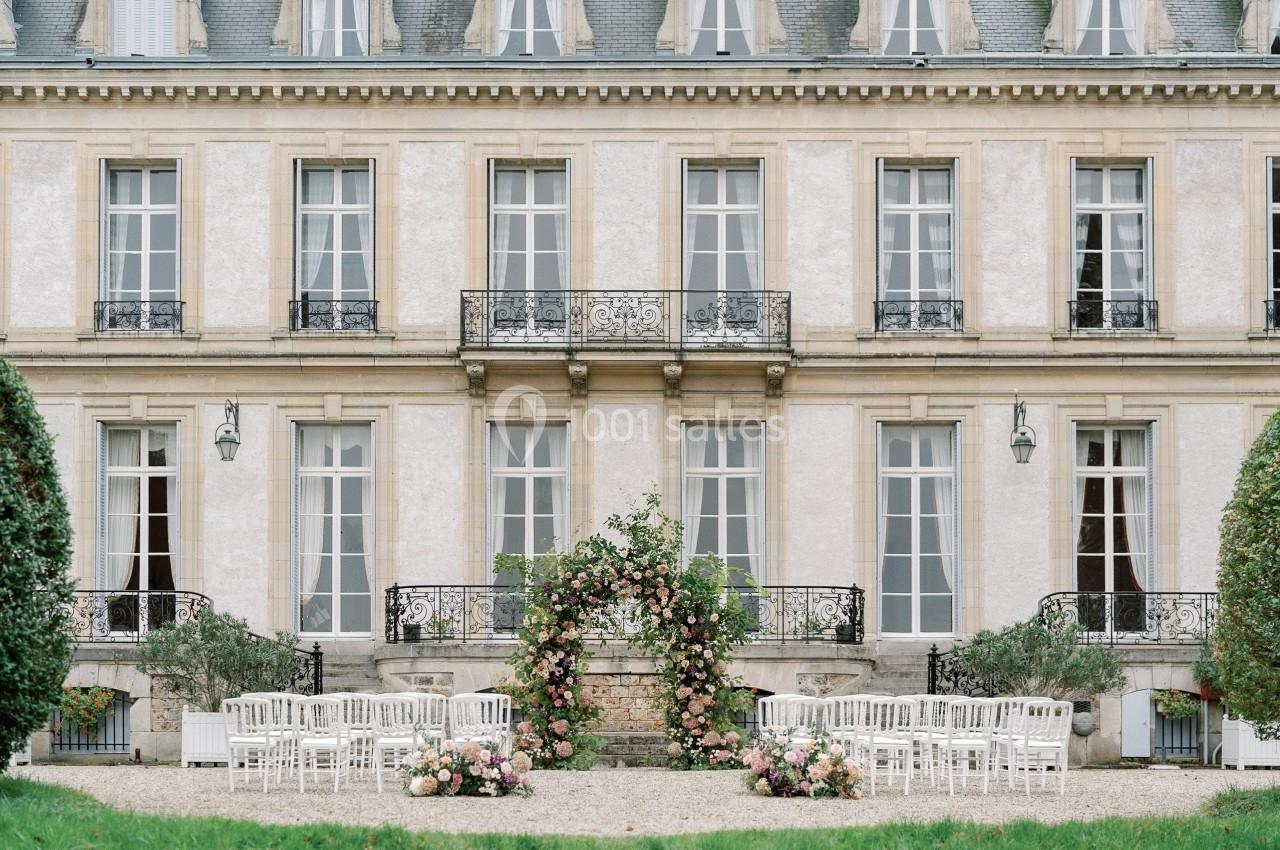Façade d'un bâtiment élégant avec des chaises blanches disposées devant une arche florale pour une cérémonie en extérieur.