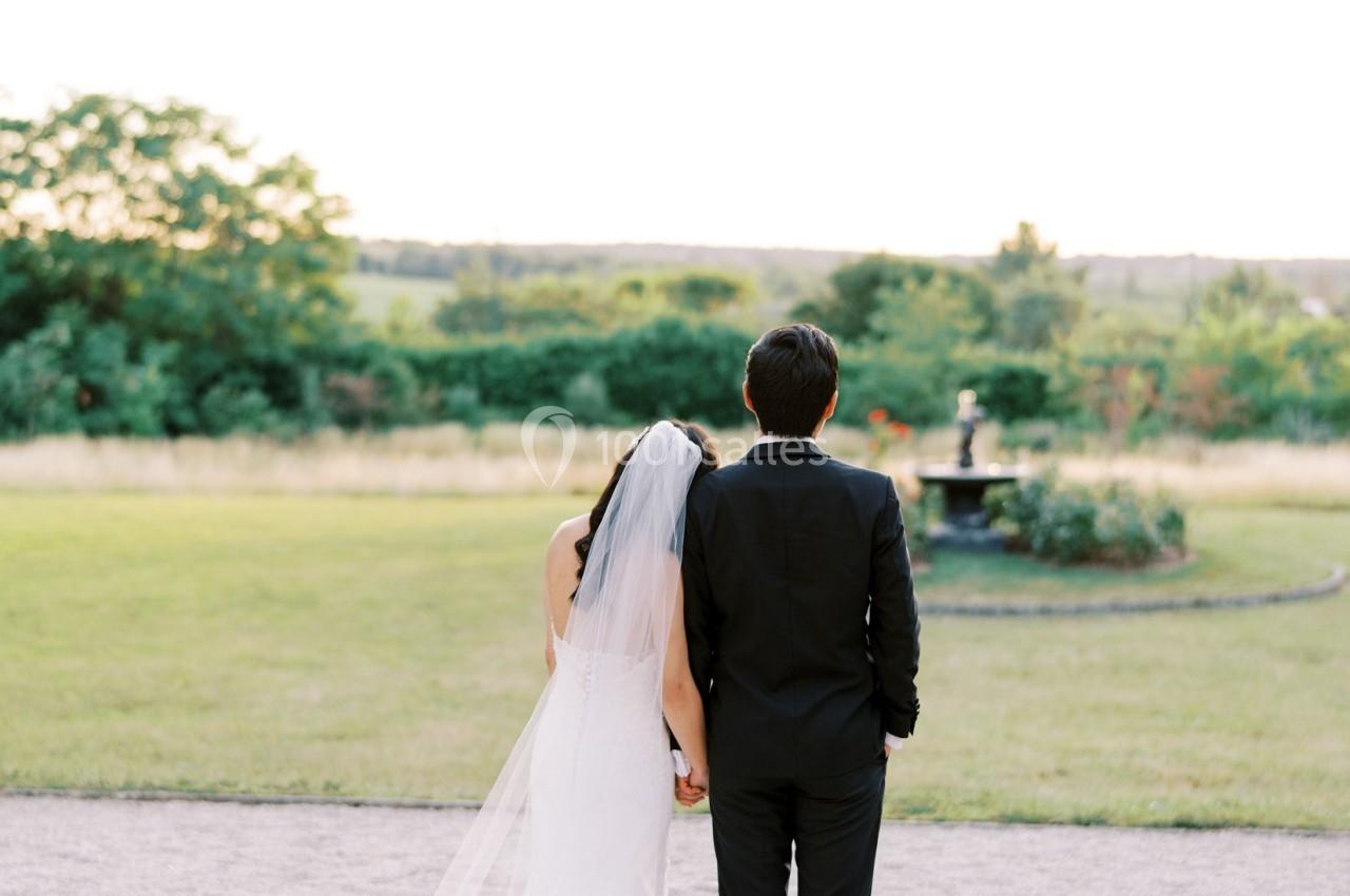 Un couple de dos, vêtu de tenue de mariage, regarde un jardin avec pelouse et fontaine sous un ciel clair.