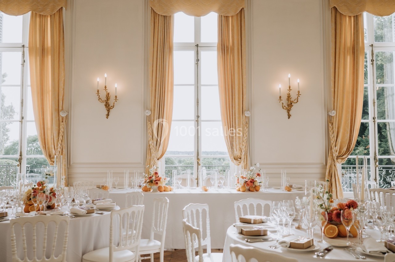 Salle de réception élégante avec tables dressées, nappes blanches, décorations florales et grandes fenêtres encadrées de…