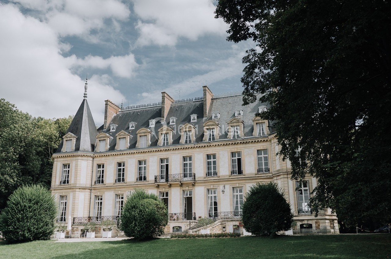 Façade d'un château historique entouré de verdure, avec un ciel partiellement nuageux en arrière-plan.