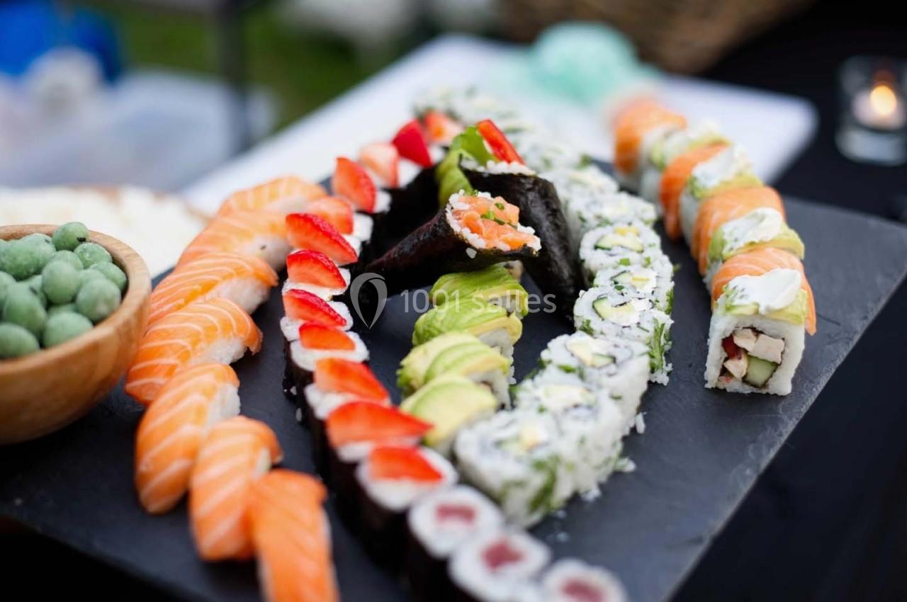 Assortiment de sushis variés disposés sur une ardoise, avec des garnitures colorées et une coupe de wasabi à côté.