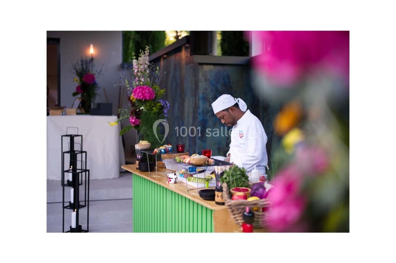 Un chef en tenue blanche prépare des plats sur un comptoir en bois, entouré de fleurs et d'éléments colorés.