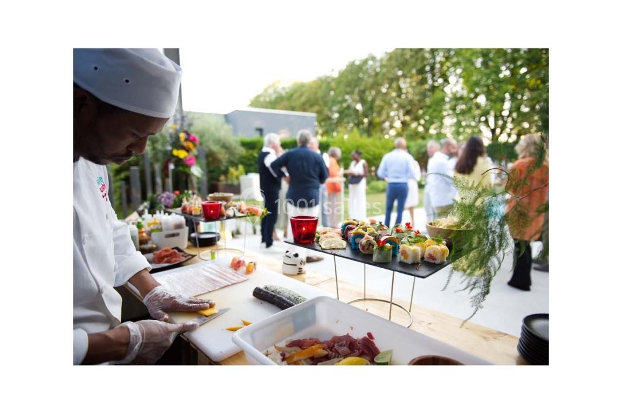 Un chef prépare des plats frais sur une table en extérieur lors d'un événement avec des invités en arrière-plan.