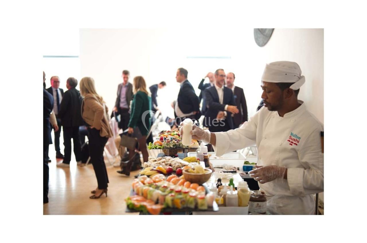 Un chef prépare des plats sur un buffet garni de fruits et boissons, avec des invités en arrière-plan.