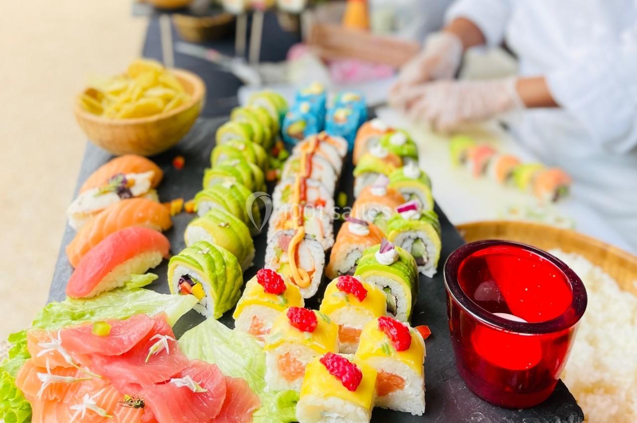 Plateau de sushis variés colorés avec un chef en arrière-plan préparant des rouleaux sur une table.