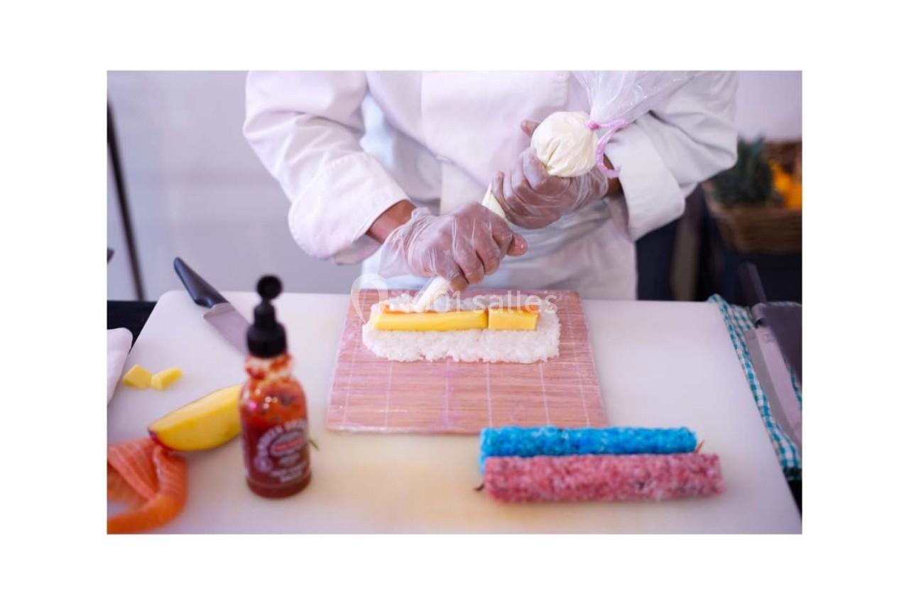 Un chef prépare un rouleau de sushi en ajoutant une garniture à l'aide d'une poche à douille sur une natte en bambou.