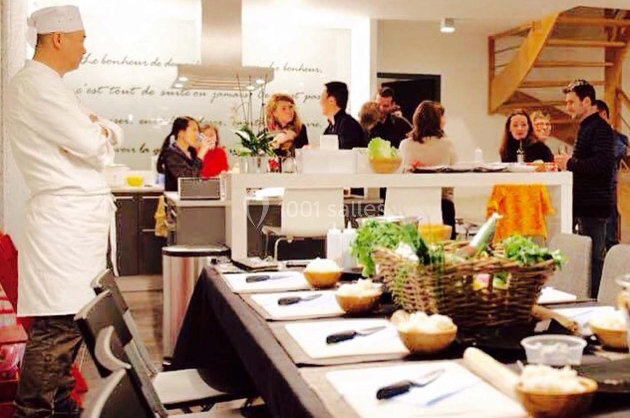 Un chef en tenue blanche observe un groupe de personnes discutant autour d'une table dans une cuisine conviviale.