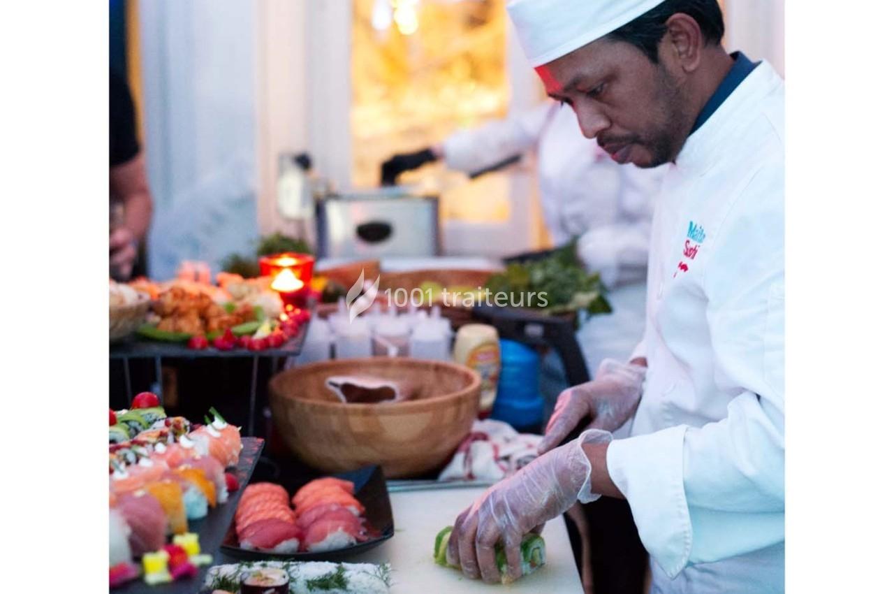 Un chef en tenue blanche prépare des sushis sur un plan de travail avec divers ingrédients et plats en arrière-plan.