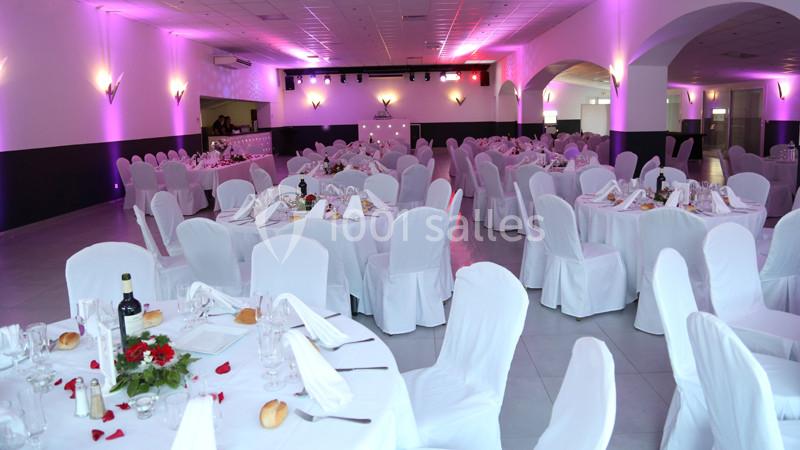 Salle de réception décorée avec des tables rondes, nappes blanches, chaises houssées et éclairage d'ambiance rose.