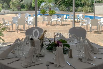 Table dressée avec nappes blanches, verres et couverts, vue sur une terrasse avec piscine et chaises en arrière-plan.
