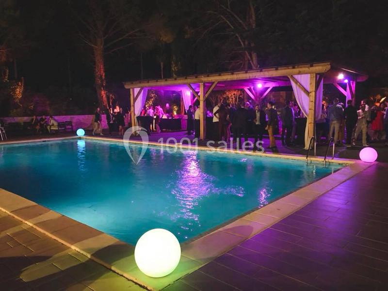 Piscine éclairée de nuit entourée de convives près d'un bar sous une pergola, ambiance festive et lumineuse.