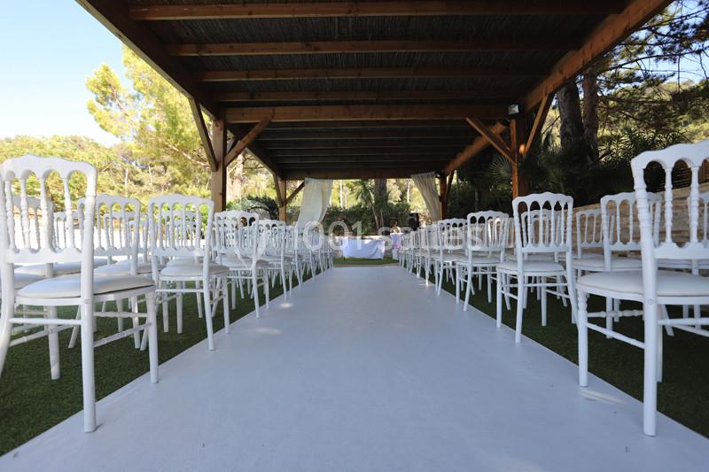 Allée centrale bordée de chaises blanches sous une pergola en bois, préparée pour une cérémonie en extérieur.
