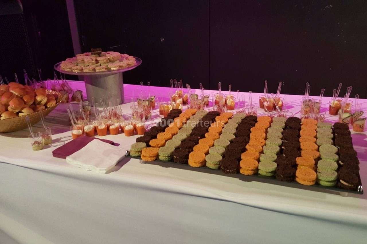 Buffet avec mini-sandwichs, verrines colorées et assortiments de bouchées sucrées et salées sur une table blanche.