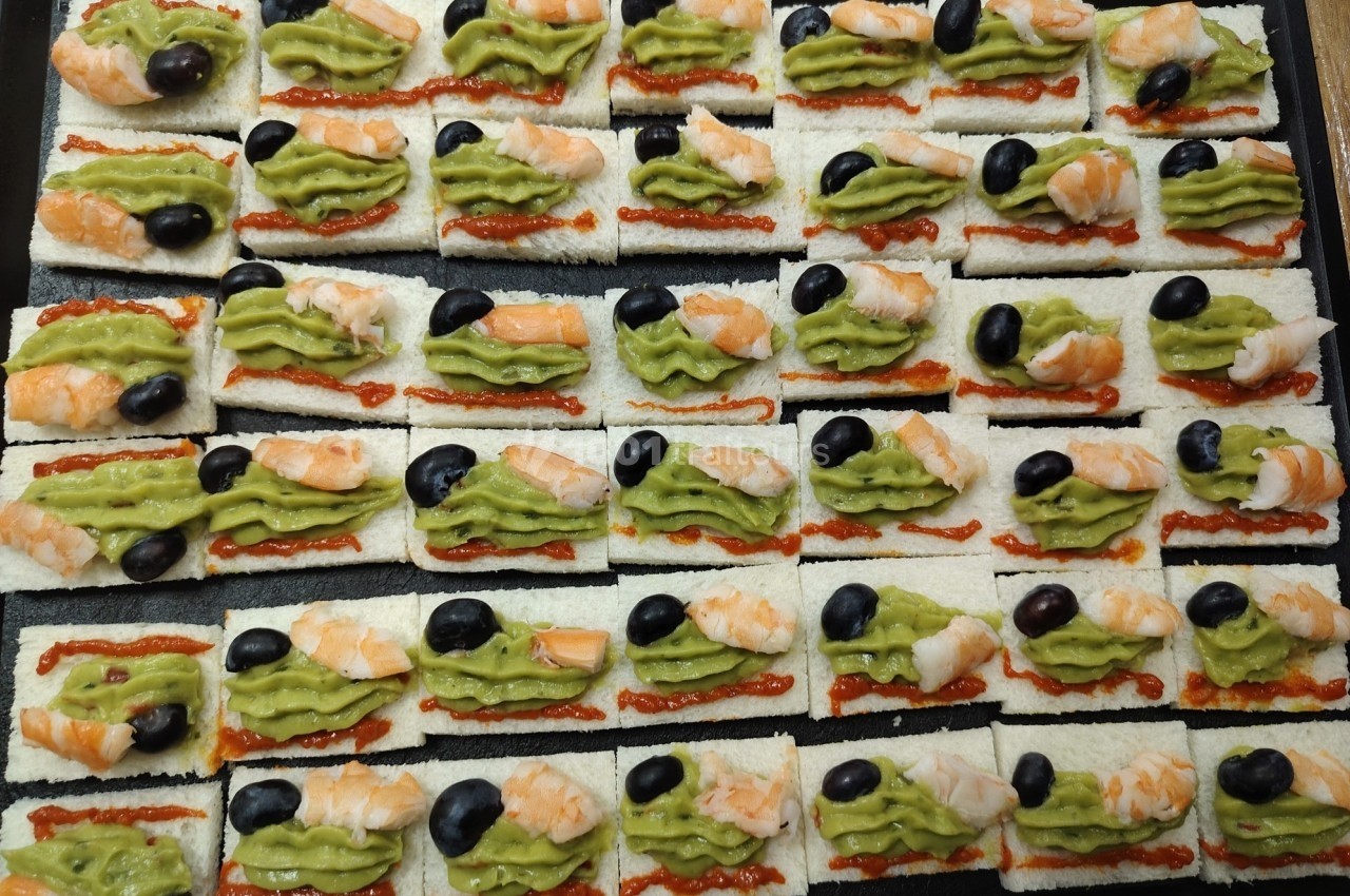 Canapés alignés avec crevettes, guacamole, olives noires et sauce rouge sur des tranches de pain blanc.