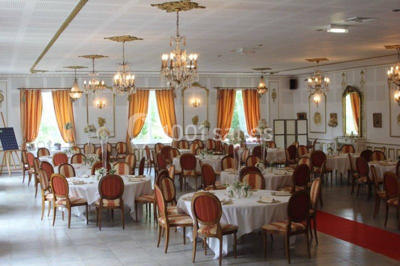 Salle de réception élégante avec tables rondes dressées, chaises rouges, lustres et rideaux orange.