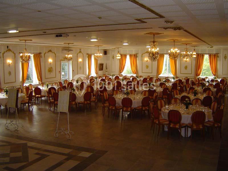 Salle de réception élégante avec tables rondes dressées, chaises rouges et rideaux orange, éclairée par des lustres.