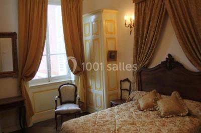 Chambre classique avec lit double, mobilier en bois, rideaux dorés et lumière naturelle entrant par une fenêtre.