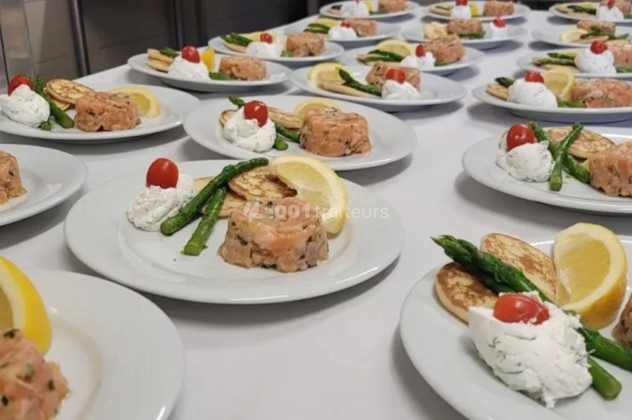 Assiettes dressées avec tartare de saumon, asperges, crème, blinis, citron et tomate cerise sur une table blanche.