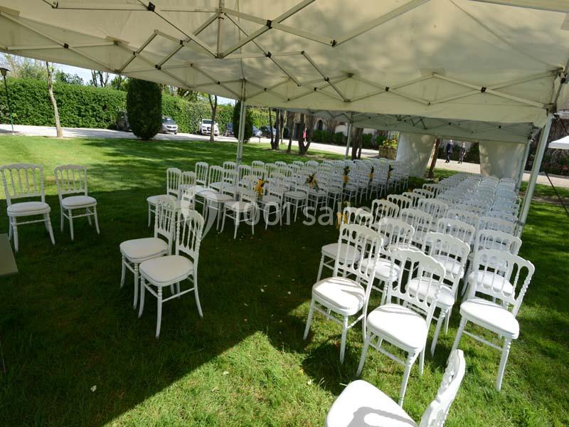 Chaises blanches disposées en rangées sous une tente dans un espace extérieur verdoyant.