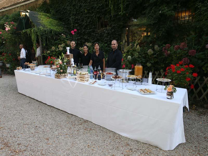 Table de buffet dressée en extérieur avec des boissons, des plats variés et quatre personnes en tenue de service.