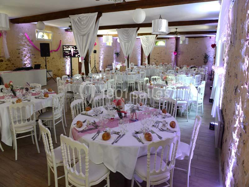 Salle de réception décorée avec des tables rondes dressées, chaises blanches et éléments de décoration rose et blanc.