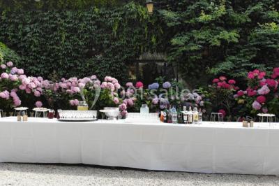 Table dressée en extérieur avec nappes blanches, boissons et verres, entourée de buissons fleuris d'hortensias.