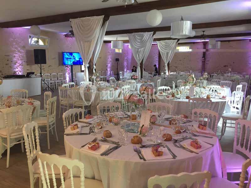 Salle de réception décorée avec des tables rondes dressées, nappes blanches, chaises élégantes et éclairage tamisé.