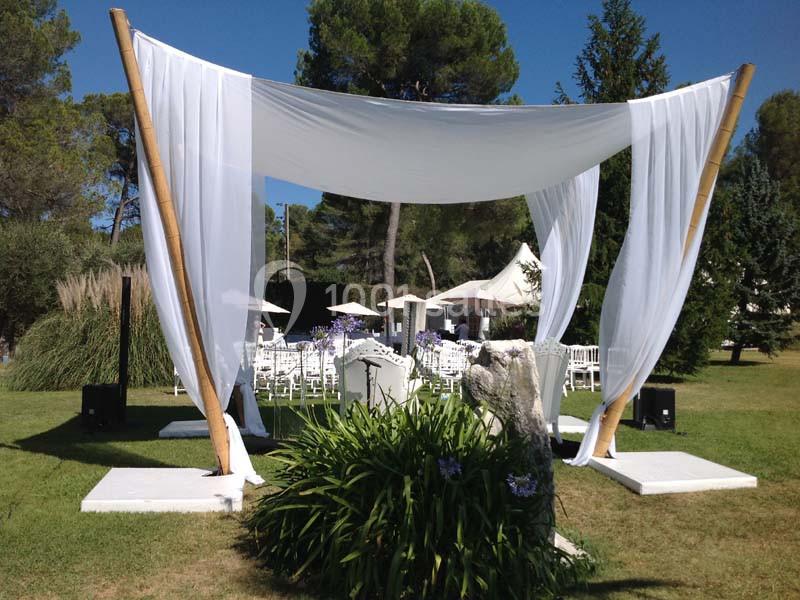 Structure de cérémonie en plein air avec voilage blanc, entourée de verdure et de chaises disposées pour les invités.