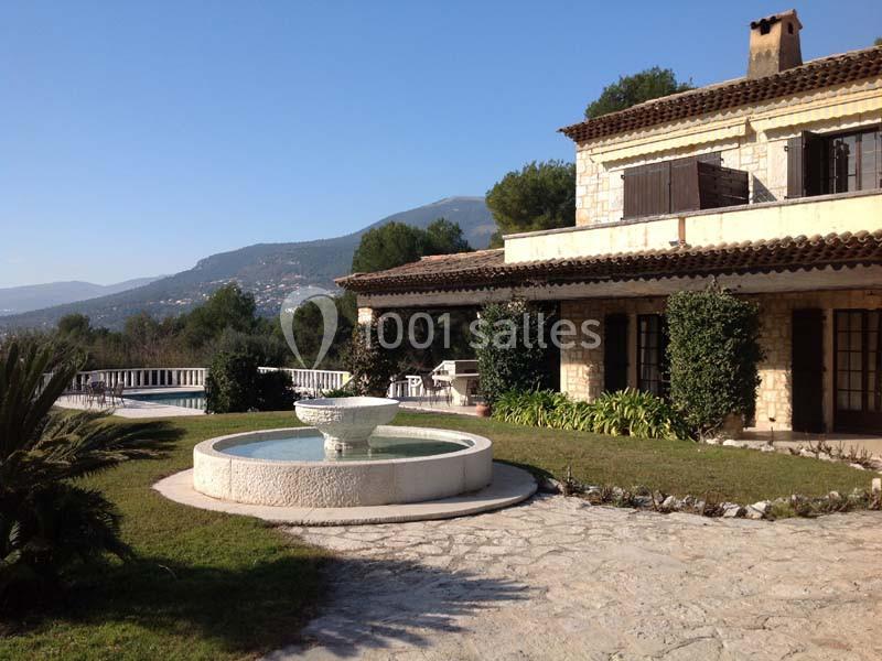 Maison en pierre avec jardin, fontaine centrale, piscine et vue sur des collines en arrière-plan.