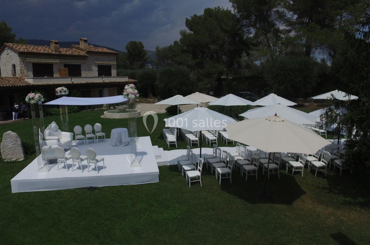 Cérémonie en plein air avec chaises blanches, parasols et décoration florale, devant une maison en pierre.