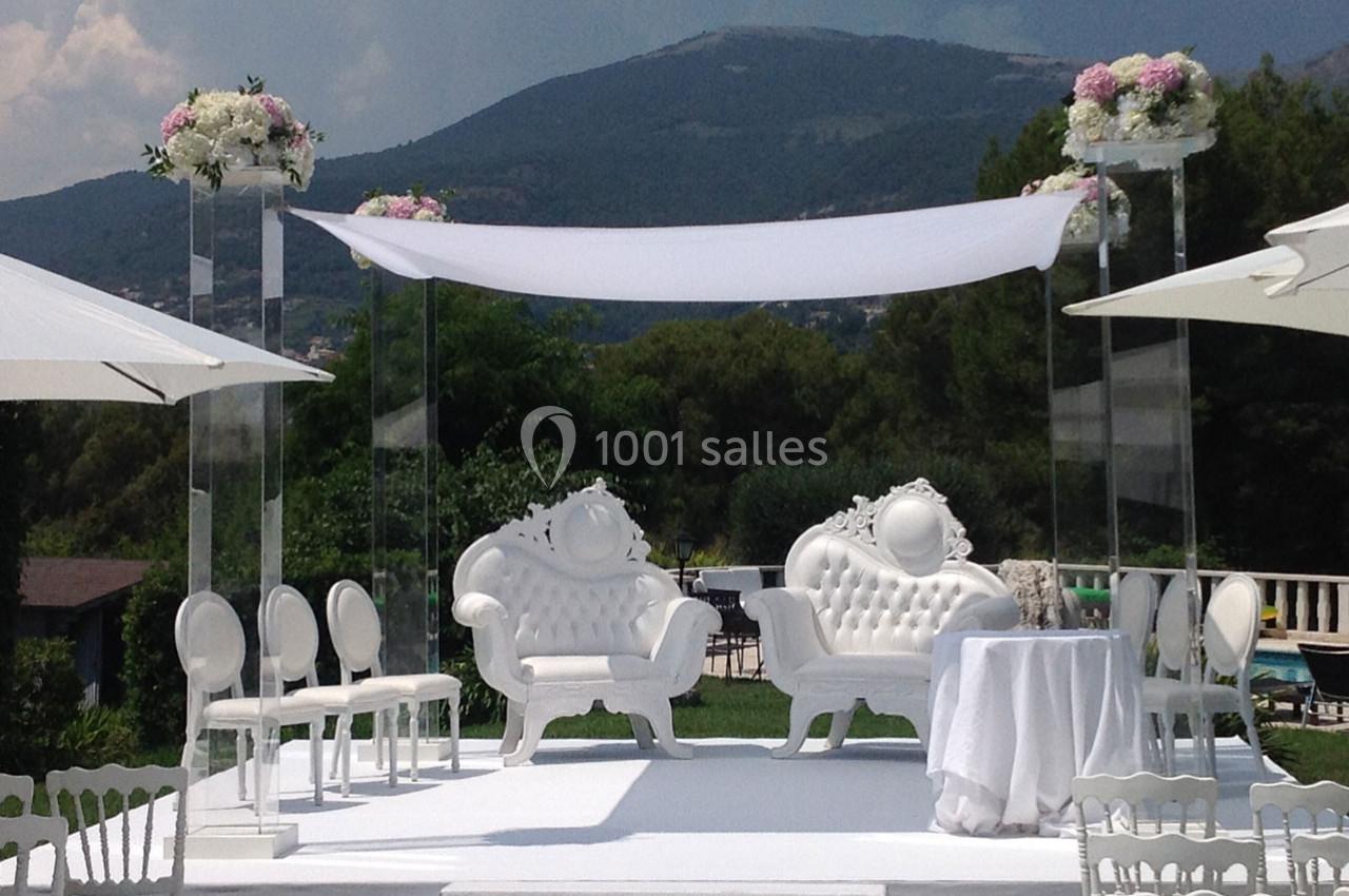 Décor de cérémonie en extérieur avec fauteuils blancs ornés, arche florale et vue sur des collines verdoyantes.