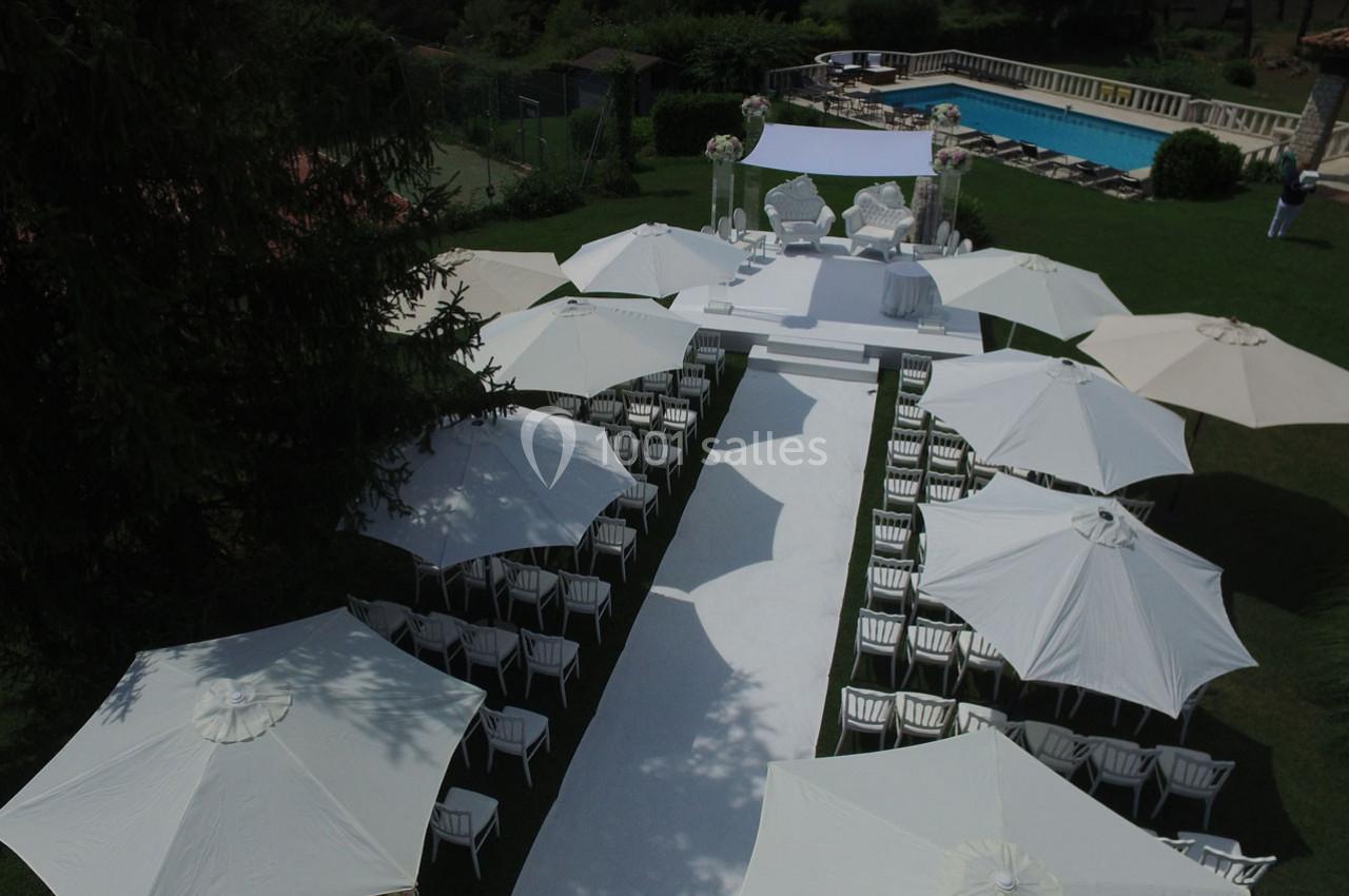 Allée centrale bordée de chaises blanches et parasols, menant à une arche décorée, dans un jardin près d'une piscine.