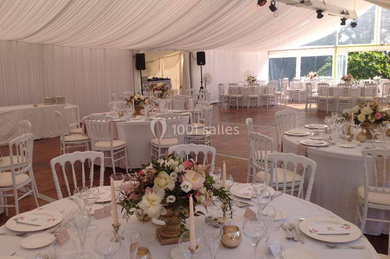 Salle de réception sous une tente blanche, tables rondes dressées avec nappes blanches et décorations florales.