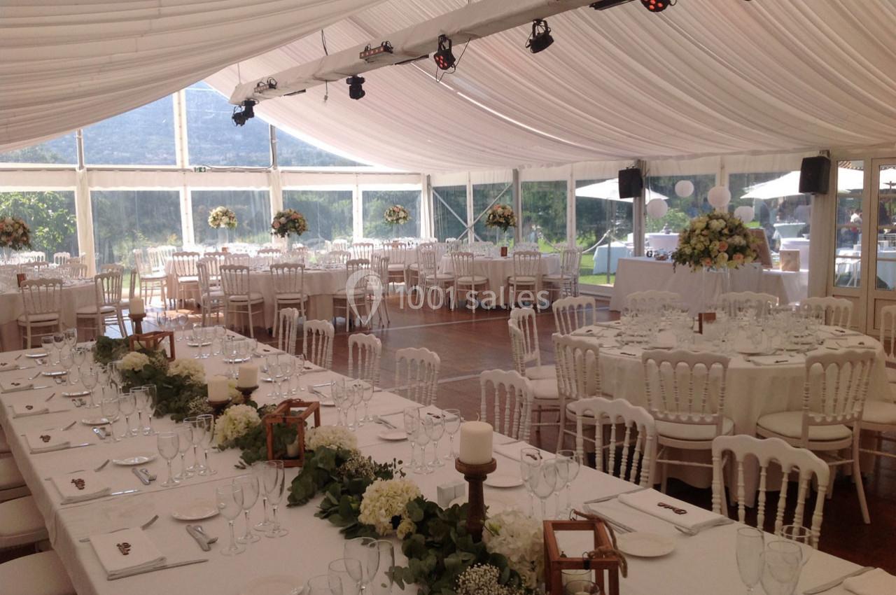 Salle de réception sous une tente blanche avec tables décorées de fleurs et chaises blanches, vue sur un jardin.