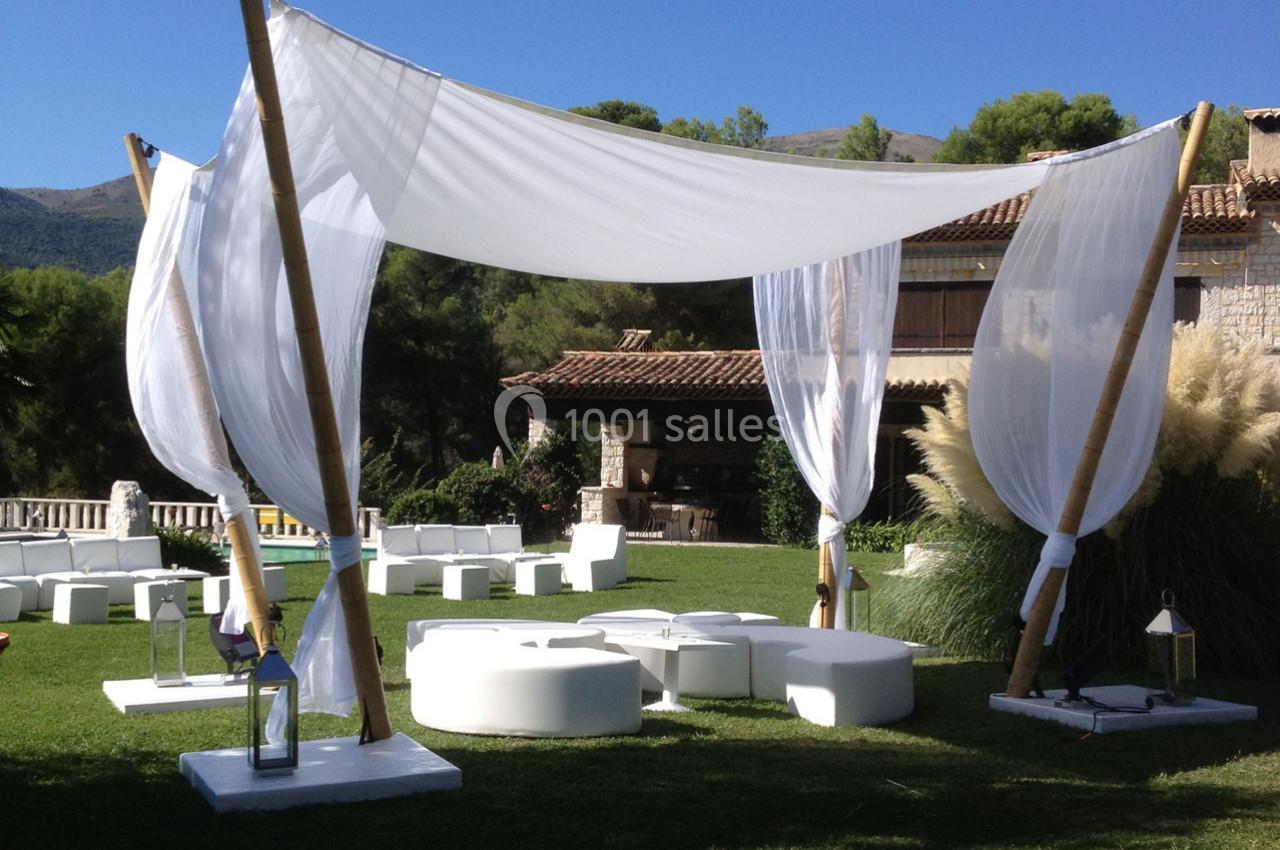 Tente légère en tissu blanc montée sur des poteaux en bois, installée sur une pelouse avec des sièges et des lanternes.
