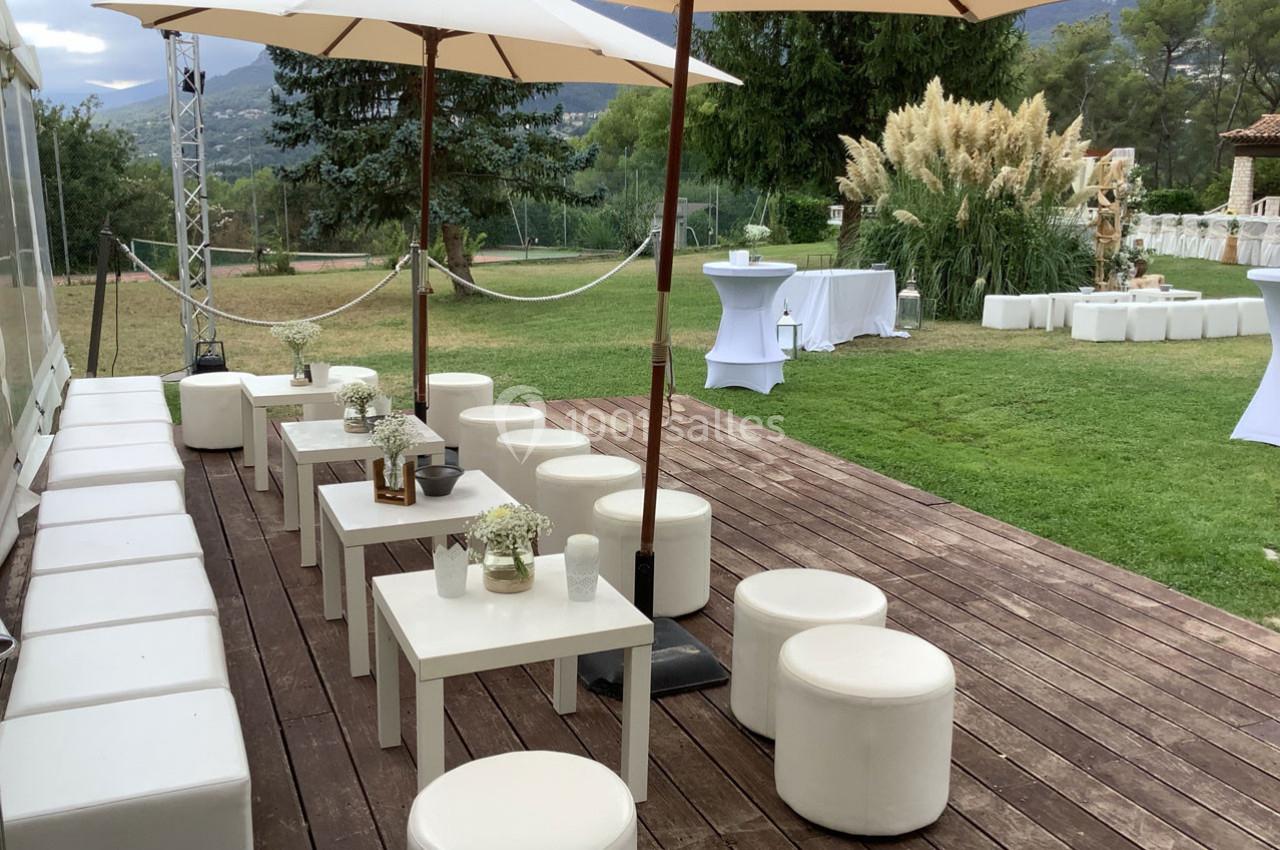 Terrasse en bois aménagée avec tables et sièges blancs, parasols et vue sur un jardin verdoyant.