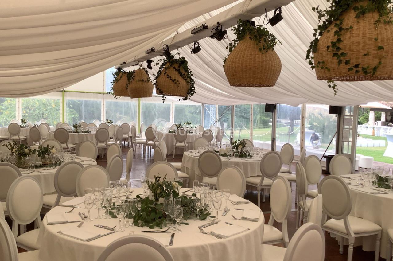 Salle de réception sous chapiteau avec tables rondes dressées, chaises blanches et suspensions en osier décorées de verdure.