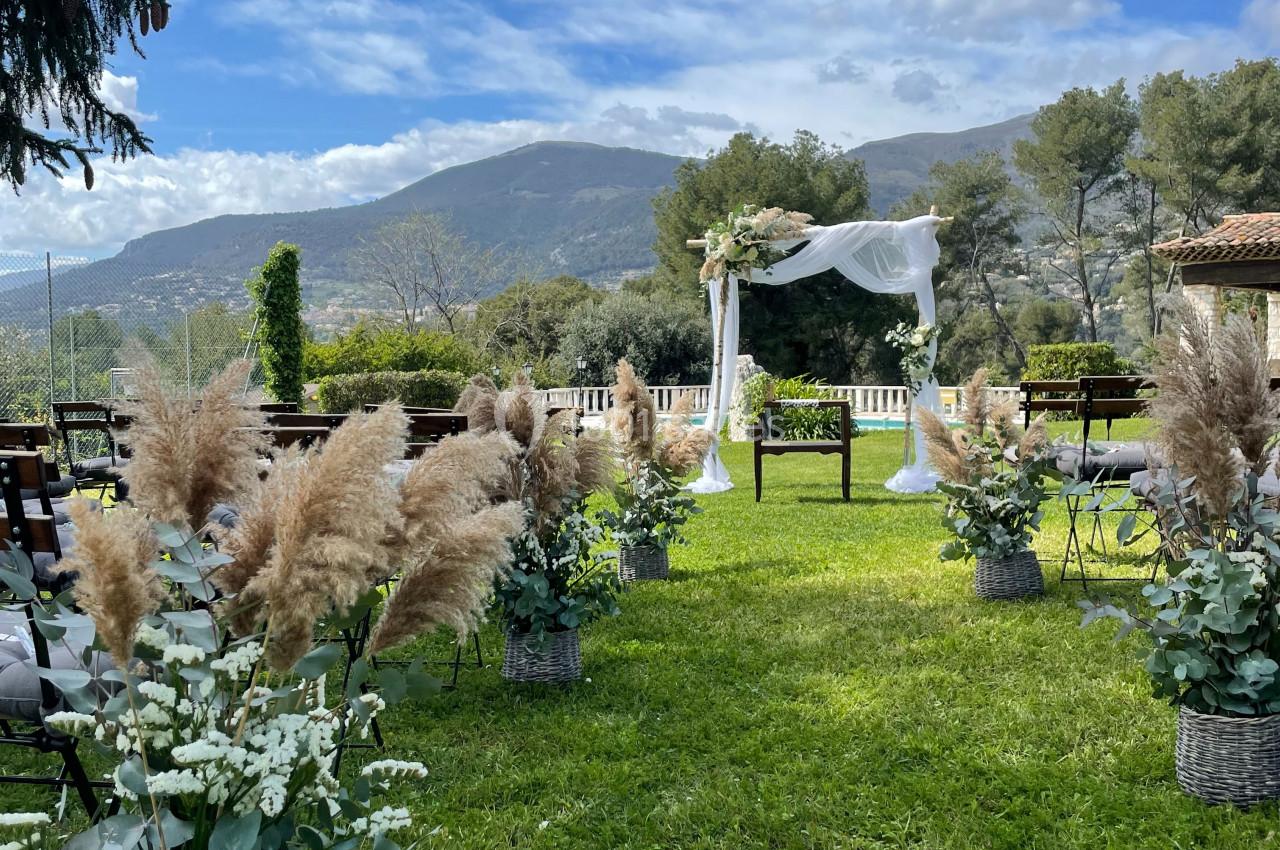 Allée décorée de fleurs et de pampas menant à une arche de mariage en extérieur, avec vue sur des collines verdoyantes.