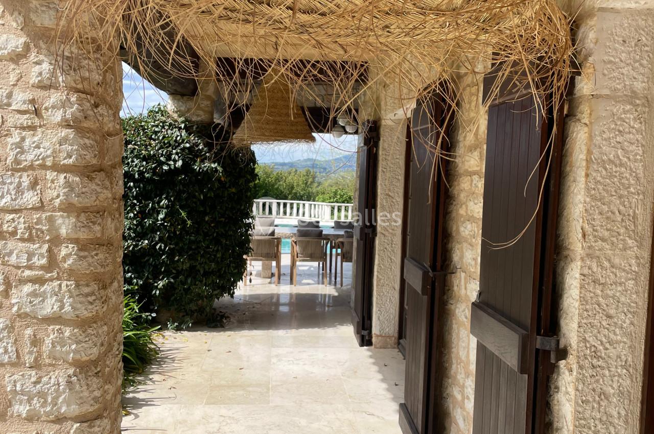 Passage extérieur en pierre avec portes en bois, grande suspension en osier et vue sur une terrasse ensoleillée.
