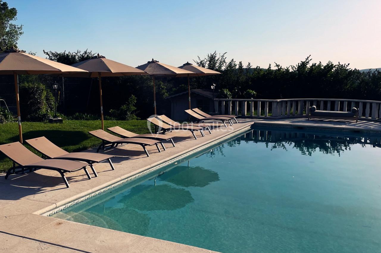 Piscine extérieure entourée de chaises longues et parasols, bordée de verdure sous un ciel dégagé.