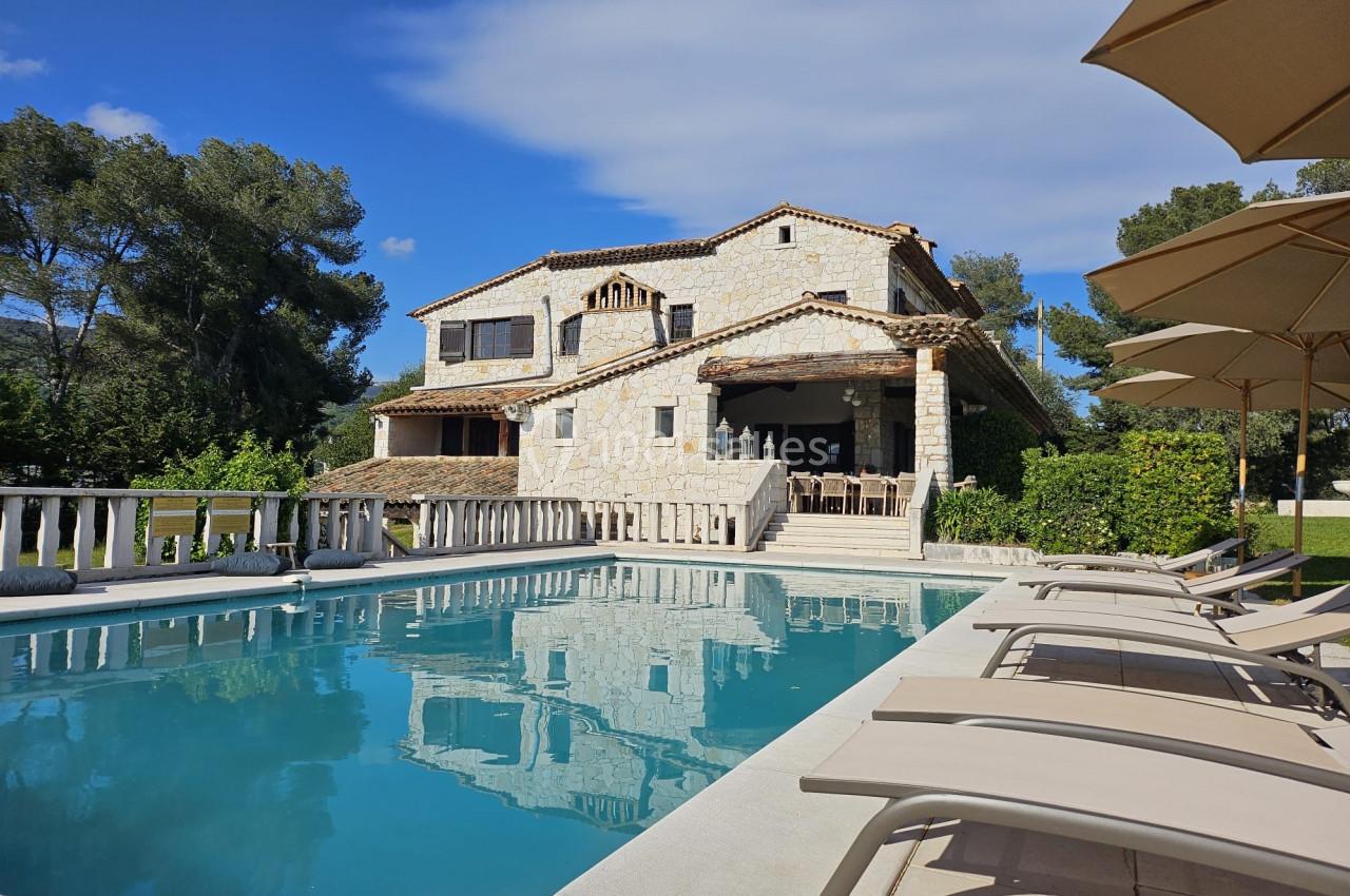Maison en pierre avec piscine, transats et parasols, entourée de végétation sous un ciel bleu.
