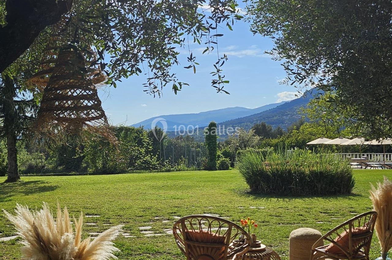 Espace extérieur avec fauteuils en rotin, tapis, plantes et vue sur un jardin verdoyant sous un ciel ensoleillé.